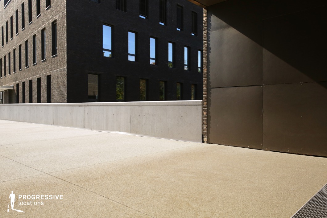 The image depicts a contemporary urban setting where industrial materials blend with clean architectural lines. A section of a building’s exterior is shown, featuring dark black brick walls contrasted by smooth concrete surfaces. The building’s geometry is sharp, with angular lines and precise edges that emphasize a modern aesthetic. A concrete barrier runs along the foreground, marking the boundary of the structure. The lighting in the image casts dynamic shadows, highlighting the materials’ textures and the sharp contrasts between the dark brick and lighter concrete. This area seems to be part of a larger architectural complex, exuding a minimalist, industrial vibe. The use of natural light adds warmth to the otherwise cool tones of the building, creating a balanced, open feel. This location is ideal for urban settings, offering a versatile backdrop for various modern, professional, or artistic projects.