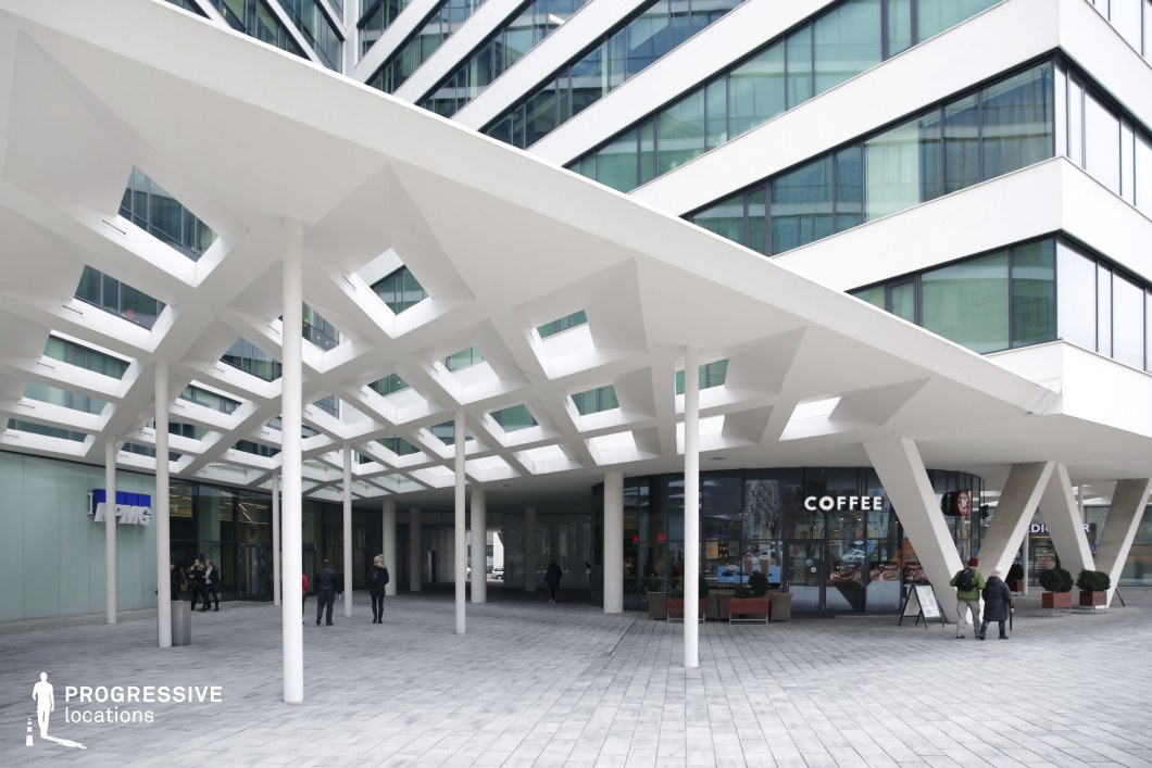 The image captures a striking modern architectural plaza, framed by angular, geometric elements. The building features a bold white facade with large glass windows, allowing natural light to flood the space. The distinctive canopy, supported by thin white columns, has a honeycomb-like structure that gives the area a unique, airy feel. Beneath the canopy, there is a spacious, paved plaza where people can be seen walking, adding life to the minimalist design. A coffee shop is visible on the ground floor, offering a welcoming atmosphere within this modern commercial complex. The wide, open layout and clean lines of the plaza provide a sophisticated, functional environment, perfect for a variety of urban activities. The overall aesthetic is sleek and professional, with a combination of contemporary architecture and urban vibrancy, ideal for business settings or social gatherings.