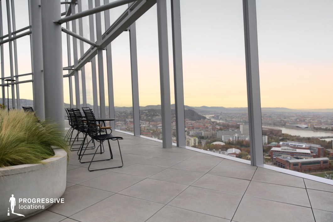 This image captures the serene and sophisticated atmosphere of a rooftop space within a skyscraper, offering a panoramic view of the city below. The large glass windows create an open, airy environment, providing an unobstructed view of the urban landscape, including the hills and river that frame the horizon. In the foreground, several black metal chairs are neatly arranged near a small table, allowing for a comfortable space to enjoy the views. A large planter with green plants adds a touch of nature to the modern architectural design. The clean, tiled flooring and steel structural supports complement the minimalist aesthetic, while the soft natural light from the fading day enhances the overall calm and inviting ambiance. This setting is ideal for high-rise office spaces or relaxation areas with sweeping views, offering a peaceful escape within a busy city environment.