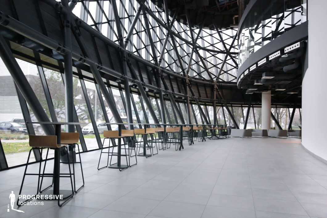 The image showcases a modern, open-plan café space with an architectural design defined by geometric steel structures and expansive glass windows. The walls are dominated by large, angled windows, allowing ample natural light to flood the interior and offering views of the outside environment. The interior features a series of sleek, high tables with wooden seating, arranged along the curved perimeter. The design is minimalist, with a neutral color palette of light gray tiles and clean lines, creating a contemporary and inviting atmosphere. The space is ideal for casual meetings or relaxation, with the exposed steel framework contributing to the industrial chic vibe. The integration of nature outside, visible through the windows, contrasts beautifully with the modern interior, making this venue suitable for urban social settings or coffee breaks in an architectural setting.