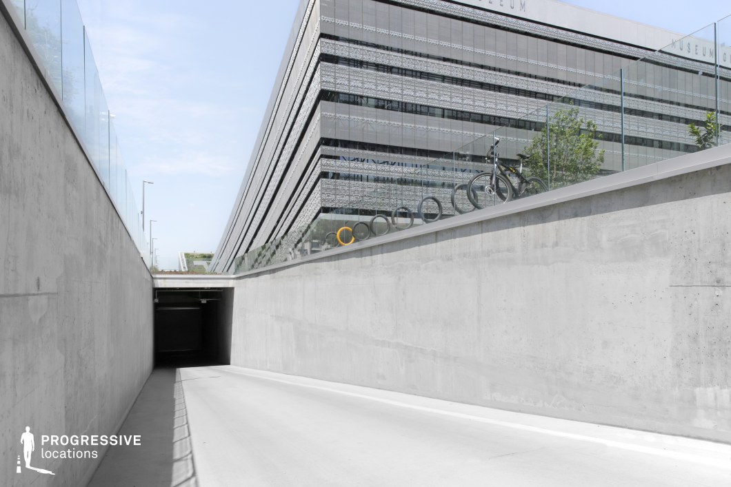 The image showcases a clean and minimalistic underground parking entrance, designed with smooth concrete surfaces and a sleek, modern aesthetic. The straight pathway leading to the tunnel emphasizes the simplicity of the space, framed by high concrete walls on either side. The upper section of the image reveals a contemporary building, with large glass windows and a distinctive architectural design, offering a contrast to the subterranean environment. The curved, sloping rooftop with green elements further enhances the modernity of the structure. Above the parking area, bike racks are neatly placed along a glass railing, adding functional detail to the setting. The clear sky and natural light contribute to the bright and open feel of the exterior, while the clean lines and industrial materials of the underground structure provide an efficient and sophisticated look. This setting is well-suited for modern urban designs, focusing on functionality with a strong emphasis on sleek, industrial aesthetics.