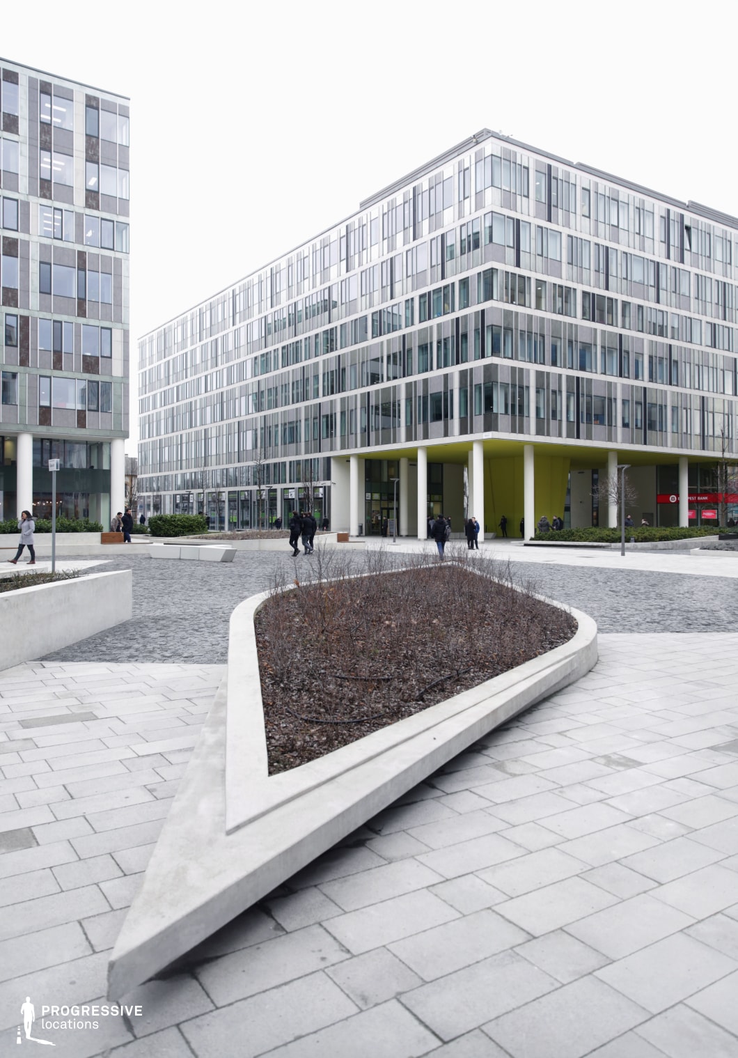 This image presents a contemporary urban courtyard framed by two modern office buildings. The structures are characterized by a sleek, uniform façade of glass and steel, creating a clean and minimalist appearance. The lower level features a spacious, open pedestrian area, with a paved surface made up of rectangular and square tiles. A distinctive, angular concrete planter, positioned in the foreground, adds a geometric element to the design, drawing attention with its sharp lines and smooth finish. The courtyard is partially landscaped with shrubs and plants, enhancing the modern yet natural feel of the space. The buildings' overhanging lower floors, supported by white columns, create a sense of openness and flow between the interior and exterior spaces. This well-maintained, aesthetically appealing area offers a versatile environment suitable for corporate settings, offering opportunities for relaxation, outdoor meetings, or casual gatherings within a clean, organized urban environment.