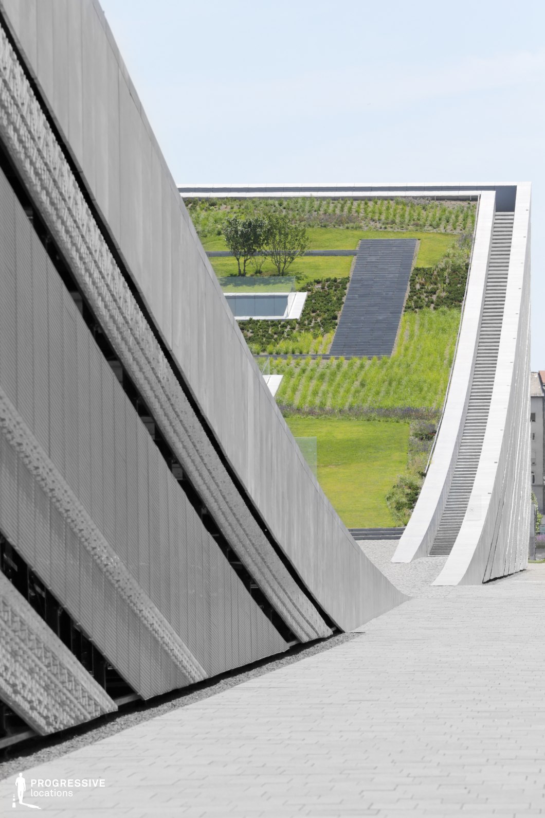 The image presents a striking architectural design characterized by sharp, sweeping curves and clean, modern lines. A large, metallic wall curves upward, leading to a stairway that ascends to a green rooftop garden. The surface of the wall is textured, with a sleek, industrial finish that contrasts with the soft, natural greenery visible at the top of the structure. The rooftop garden is meticulously landscaped with neatly arranged grass and trees, creating a harmonious blend of urban architecture and nature. The curved path and stairs guide the eye upward, enhancing the sense of movement and dynamic space. This design emphasizes a modern, minimalist aesthetic while incorporating natural elements, making it ideal for architectural projects that highlight sustainability and urban integration. The combination of angular concrete surfaces and the lush rooftop garden creates an intriguing visual contrast, suitable for high-end urban developments or public spaces.