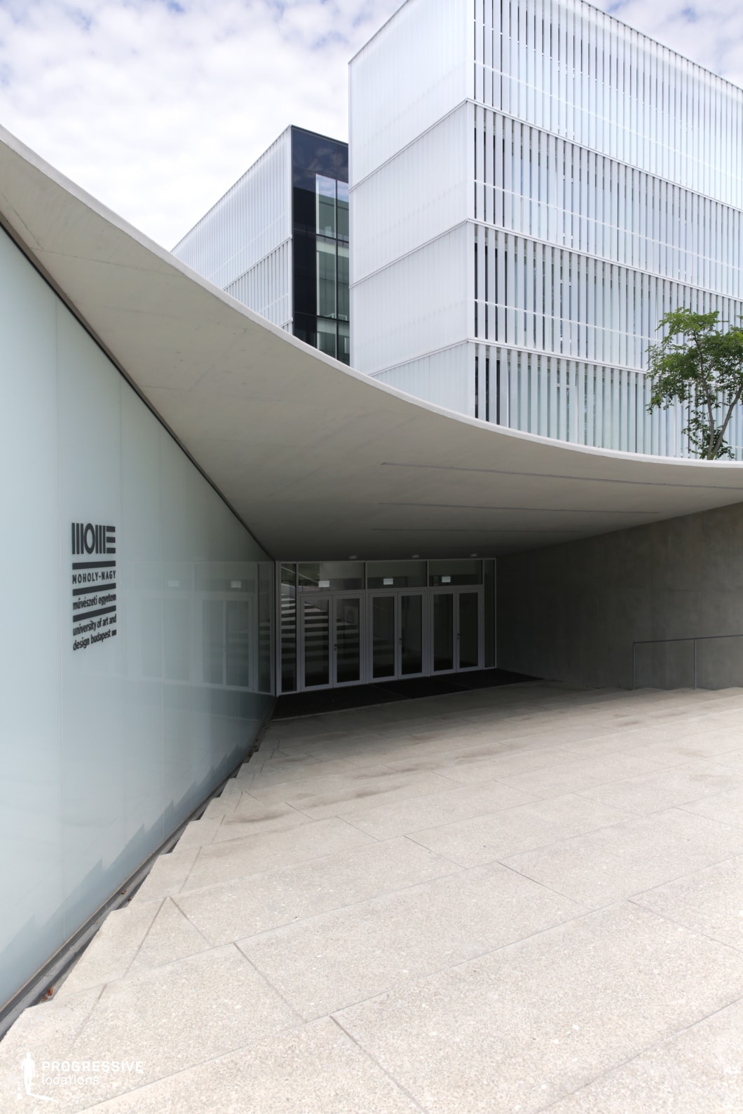 The image captures the sleek and modern entrance of a university building, characterized by a smooth, curved canopy that extends over the entrance area. The canopy’s gentle slope contrasts beautifully with the sharp, angular edges of the surrounding glass and concrete structures. The façade is made of large glass panels that allow for a clear view into the building, while the concrete ground is paved with large, smooth tiles, creating a clean and minimalist look. The stairway leading up to the entrance is subtly integrated into the design, with a handrail running along the right side for accessibility. The overall aesthetic is modern, with an emphasis on simplicity and functionality. The building's architectural elements highlight the contemporary design trends in educational spaces, combining both open, welcoming features with strong geometric lines. This space is ideal for projects requiring a dynamic yet refined atmosphere, suitable for academic or professional settings.