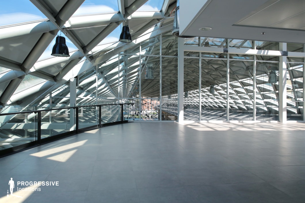 The image showcases a modern event hall characterized by its expansive glass façade and dynamic metal roof structure. The ceiling features a distinct, angular metal framework that creates a visual connection with the architectural design, resembling the ribs of a whale. This geometric framework adds depth and intrigue to the space, while the large windows provide an abundance of natural light, further highlighting the intricate design. The floor is composed of large, light-colored tiles, contributing to the clean, open atmosphere. The space appears vast and versatile, ideal for events, exhibitions, or gatherings, with the unique architectural features providing a stunning backdrop. The interplay of light and shadow across the room enhances the visual appeal, creating an atmosphere of sophistication and modernity. The design emphasizes a balance of structure and openness, making it suitable for a wide range of creative and professional projects.