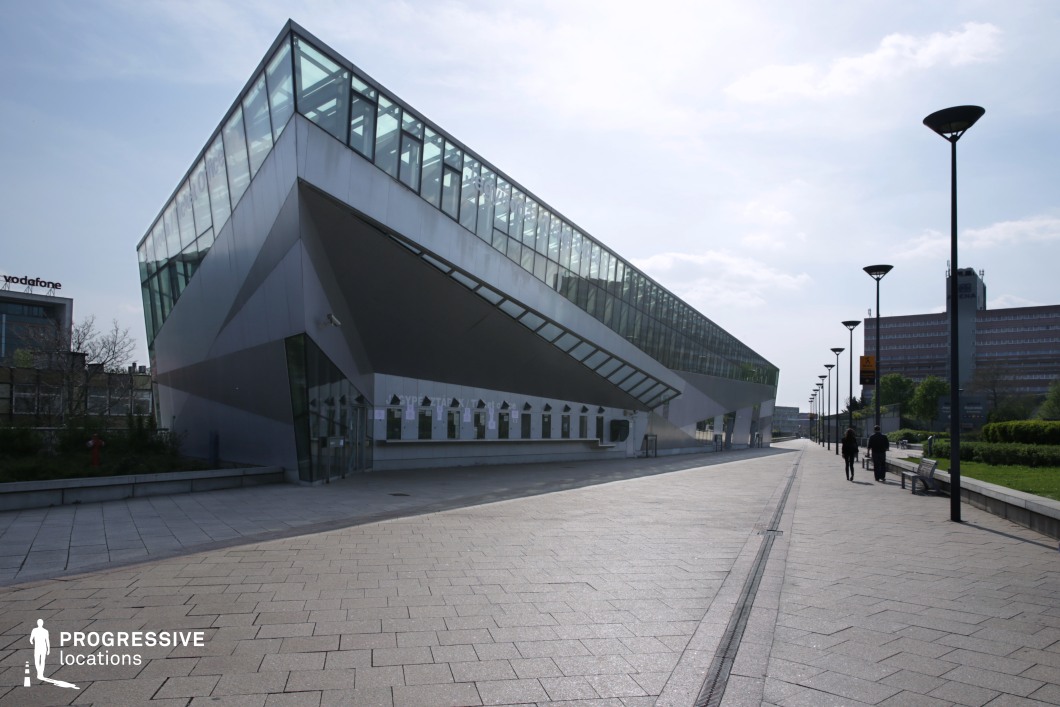 This image showcases a contemporary event hall with bold, angular architectural elements that create a strong visual impact. The building features a sharply slanted roof, giving it a dynamic, futuristic feel. The glass facade on the upper portion of the structure is sleek, offering an open and airy atmosphere that contrasts with the solid, geometric lower half. The exterior is finished with smooth, metallic surfaces that reflect light, adding to the building's modern aesthetic. A set of ticket office windows is located at the base, providing access to the hall, while the large glass windows above enhance the building’s open, inviting appearance. The surrounding area is neatly paved, with streetlights lining the path and benches offering seating along the walkway. The combination of clean lines, reflective surfaces, and well-designed landscaping makes this location ideal for dynamic, high-end event settings. This space would be excellent for contemporary film production scenes, providing a visually compelling backdrop that is both modern and functional. The open architecture allows for dramatic shots, while the geometric angles and lighting could be used creatively to emphasize action or add a cinematic flair.