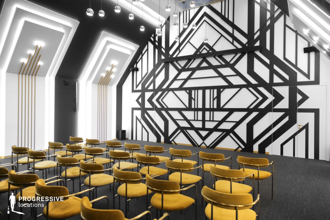 This image showcases a striking conference room designed with bold geometric patterns and a modern aesthetic. The room is dominated by a large, black-and-white cubist mural on the back wall, featuring sharp angles and intersecting lines that create a dynamic, visually stimulating effect. The ceiling is adorned with sleek, contemporary lighting, and the walls feature gold-accented vertical lines, adding warmth to the otherwise minimalist color palette. The room is filled with neatly arranged yellow chairs that contrast beautifully with the black and white tones, creating a balance of color and style. The geometric design and lighting emphasize the modernity and creativity of the space, making it an ideal location for creative meetings, presentations, or artistic events. The combination of geometric patterns and functional furniture creates a space that is not only visually engaging but also practical for various professional activities, offering a contemporary and stylish backdrop for filming or photography.