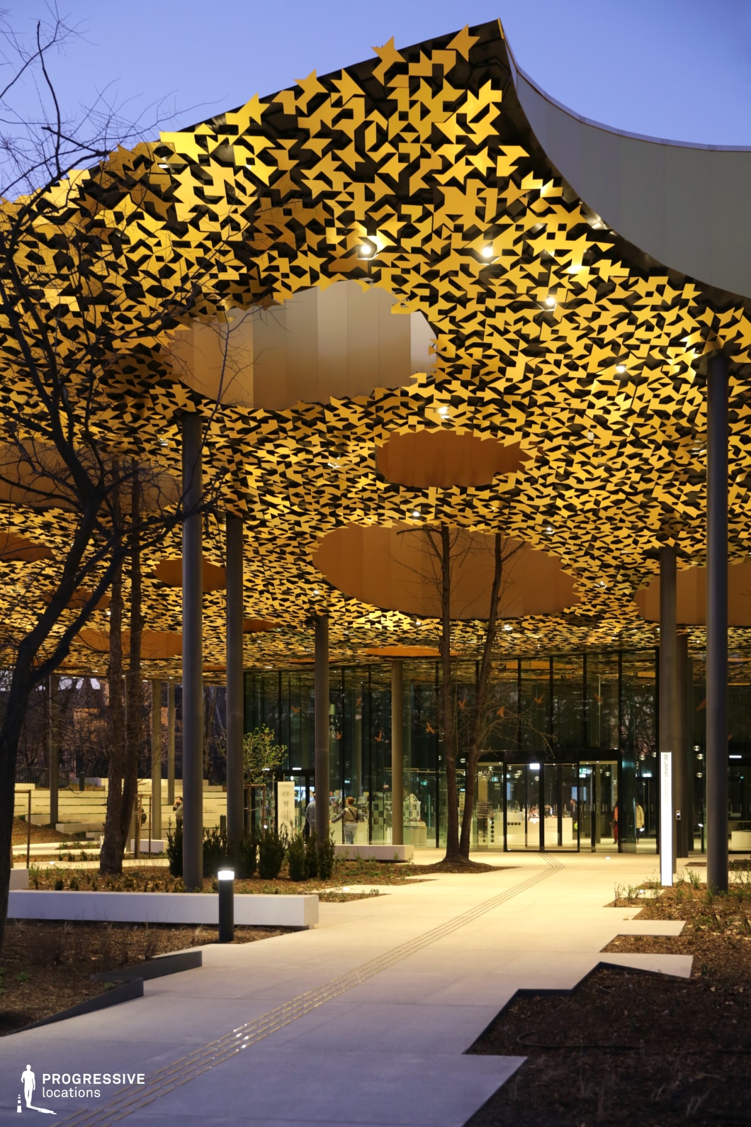  This image captures a stunning concert hall entrance, highlighted by a dramatic canopy design. The roof is adorned with intricate golden patterns, resembling abstract leaves or geometric shapes, which create a striking visual effect against the twilight sky. The golden canopy is suspended above tall, sleek columns, adding an element of elegance and grandeur to the space. Beneath the canopy, the modern glass facade of the building offers a glimpse of the interior, showcasing the transparent, open design. The surrounding plaza is well-lit, with trees and neatly arranged landscaping contributing to the welcoming atmosphere. The combination of the golden, ornate ceiling and the contemporary architecture creates a visually compelling and dynamic space, ideal for performance or cultural events. This location’s blend of artistry and modernity makes it an excellent choice for filming high-profile events, providing both an atmospheric exterior and a visually captivating focal point for dramatic shots. The elegant lighting and intricate design would work well in films that require sophisticated or artistic settings.