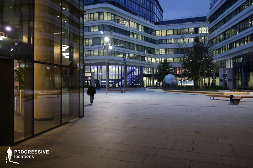 This image features a contemporary office building complex, illuminated at night to highlight its sleek glass façade and modern design. The building’s curved, glass exterior reflects the soft glow from the outdoor lights, emphasizing the clean, minimalist architecture. The well-lit courtyard between the structures contains a large spherical sculpture and rows of benches, offering a peaceful, open space amidst the bustling urban environment. Streetlights and subtle lighting along the pathways add to the atmosphere, enhancing the modernity of the space. The overall design combines functionality and aesthetics, creating a visually appealing environment ideal for corporate offices, hotel lobbies, or urban filming. The interplay of light, glass, and clean lines would work well in high-end commercial film productions or urban architectural photography, offering a polished and sophisticated backdrop for various creative projects.