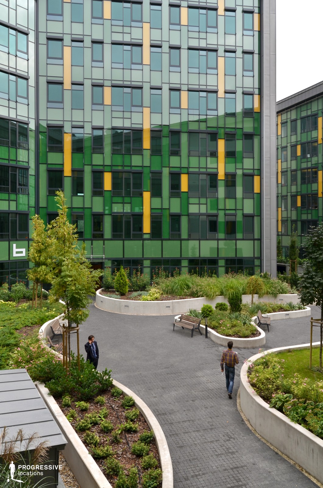 This image depicts a vibrant, modern office complex surrounded by a beautifully landscaped garden. The building’s facade features a striking pattern of green and yellow panels that give it a dynamic and contemporary look. Large glass windows complement the colorful design, providing ample natural light and reflecting the lush garden below. In the foreground, a well-maintained garden with curved concrete planters adds to the tranquil atmosphere, featuring neatly arranged benches for relaxation. The walkway, composed of dark paving stones, winds through the garden, providing a path for people to walk through the space. The overall environment blends nature with modern architecture, creating a welcoming space ideal for work or social interaction. This location is perfect for film production, offering a mix of architectural interest, greenery, and a lively yet peaceful atmosphere, making it suitable for scenes that emphasize modern, urban life or outdoor business environments.