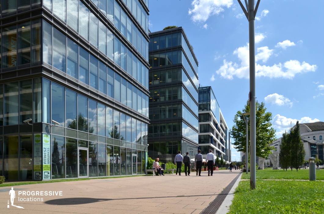 This image showcases a vibrant, modern business promenade set against the backdrop of sleek glass office buildings. The well-maintained walkway is lined with green spaces and decorative trees, contributing to a clean and professional atmosphere. The buildings themselves are characterized by large glass panels, reflecting the sky and surrounding environment, while the expansive windows give a sense of openness and transparency. Benches provide areas for relaxation, complementing the modern architecture and the natural elements that surround the area. The bright sunlight and clear blue skies enhance the dynamic feel of the space, making it ideal for showcasing corporate environments, modern work settings, or contemporary urban living. The space’s blend of sleek architecture and greenery provides an excellent backdrop for filming business, lifestyle, or commercial scenes, creating an upscale, urban aesthetic.