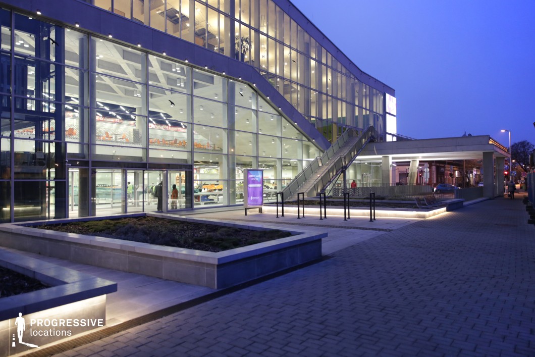 This image captures the exterior of a modern building at dusk, showcasing its sleek, glass-fronted architecture. The building features large windows that allow an interior view of the space, with visible bright lights from within. A prominent staircase on the right side of the image leads up to the building's entrance, adding depth to the structure’s design. The surrounding plaza is beautifully lit by soft lighting along the walkways, accentuating the contemporary feel of the environment. A small landscaped area with low lighting complements the overall aesthetic, enhancing the sleek, urban feel of the scene. The twilight sky, combined with the building's bright interior and exterior lights, creates a dynamic contrast, highlighting the building's modern features. This setting, with its clean lines and atmospheric lighting, offers a versatile backdrop ideal for filming nighttime urban scenes or business-related activities. The architectural layout and lighting design make it suitable for high-end commercial and architectural photography or film productions.