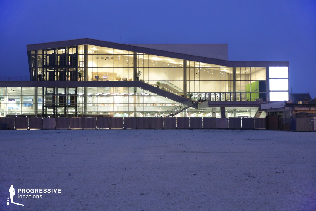 This image showcases a modern building illuminated at night, featuring a striking glass façade that allows a clear view of the interior. The structure’s unique stepped design adds a dynamic and sculptural element to its appearance, with wide staircases leading up to the entrance. The illuminated interior creates a bright, welcoming atmosphere, with soft lighting highlighting the architecture and spaces within. The building's contemporary design, with its clean lines and expansive glass surfaces, is set against a barren landscape, which adds to its prominence. This location provides an excellent backdrop for filming modern corporate, commercial, or artistic scenes, offering a sophisticated and visually compelling setting. The combination of the building's modern design and the soft nighttime lighting creates an ideal atmosphere for both exterior and interior shots in film and photography.