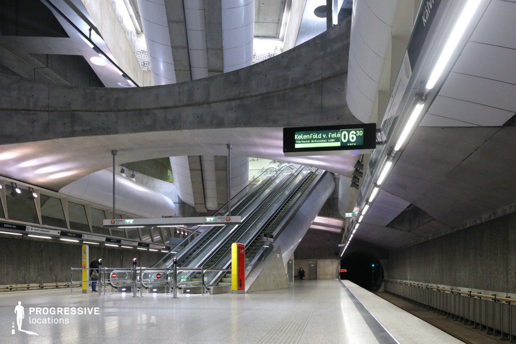 This image presents a futuristic and minimalist metro station with a spacious platform, characterized by sleek concrete surfaces and angular architecture. The lighting system is efficiently integrated into the ceiling, highlighting the clean lines and industrial design elements of the station. The escalators lead upwards, creating an active yet visually balanced composition. Information signage displays the next train schedule, adding a functional, urban touch. The environment is illuminated with soft artificial lighting, creating a warm contrast against the cool tones of the concrete and steel structure. The space is ideal for filming urban, action-packed scenes or modern city-based scenarios, offering a dynamic and atmospheric setting. The minimalist design and modern functionality of the metro station provide versatility for various film genres, particularly in creating scenes that evoke a contemporary, high-tech urban environment.