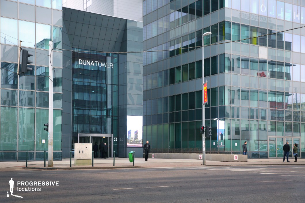 This image showcases a sleek, modern office building with a reflective glass façade, creating a striking visual effect as it mirrors its surroundings and the sky. The large windows allow for an abundance of natural light, complementing the contemporary design. The entrance is marked by a clean, minimalist approach with well-lit glass doors. The surrounding environment includes modern street infrastructure, including traffic lights and public signage, contributing to the urban setting. The building's glass and steel structure creates a futuristic, minimalist ambiance, making it ideal for high-tech corporate scenes or city life depictions in films.