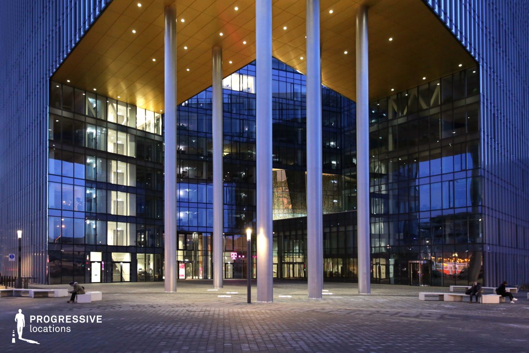 This modern office building features striking, glass-walled exteriors illuminated from within, creating an attractive and inviting atmosphere at night. The structure is supported by a series of prominent metallic pillars that form a distinct entrance canopy, showcasing a blend of contemporary architectural design and functional form. The building's sleek lines and reflective glass surfaces add sophistication, while the well-lit plaza area in front enhances the space's urban charm. The open space around the pillars and building's base is ideal for capturing a clean, professional scene, perfect for portraying business environments, urban settings, or modern corporate locations in film production. The lighting at night adds drama and depth, creating a visually dynamic scene for exterior shots.