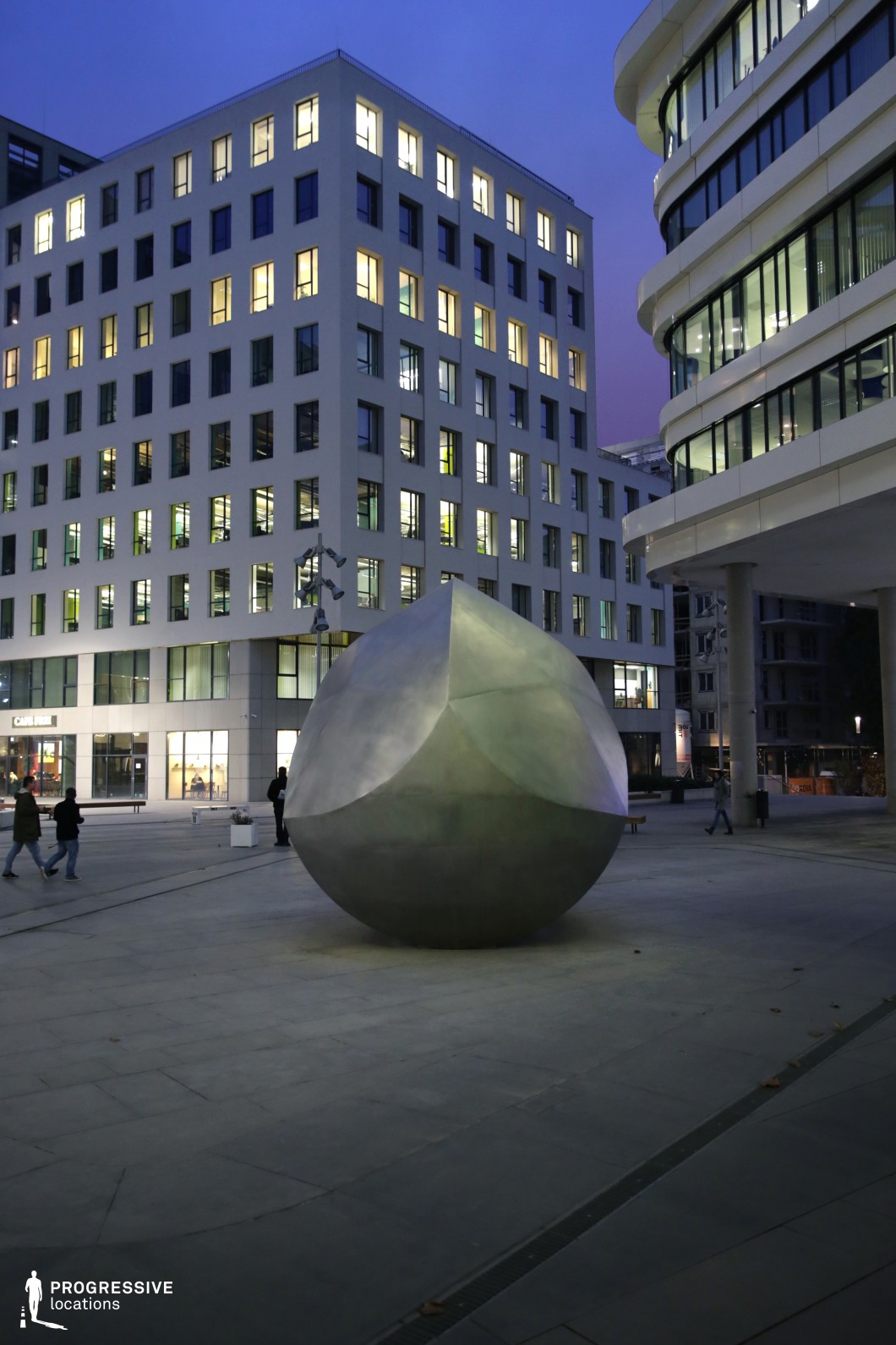 The photograph features a sleek and modern office plaza with a striking metallic orb sculpture positioned at the center of the open space. The surrounding office buildings, characterized by geometric design and large windows, provide a clean, contemporary backdrop. The windows emit warm light, adding a sophisticated atmosphere to the setting. This location is ideal for filming urban scenes, corporate settings, or contemporary lifestyle narratives, with its minimalistic architecture and ample space for creative shots. The design elements make it a perfect setting for conveying a polished, business-oriented environment in film productions.