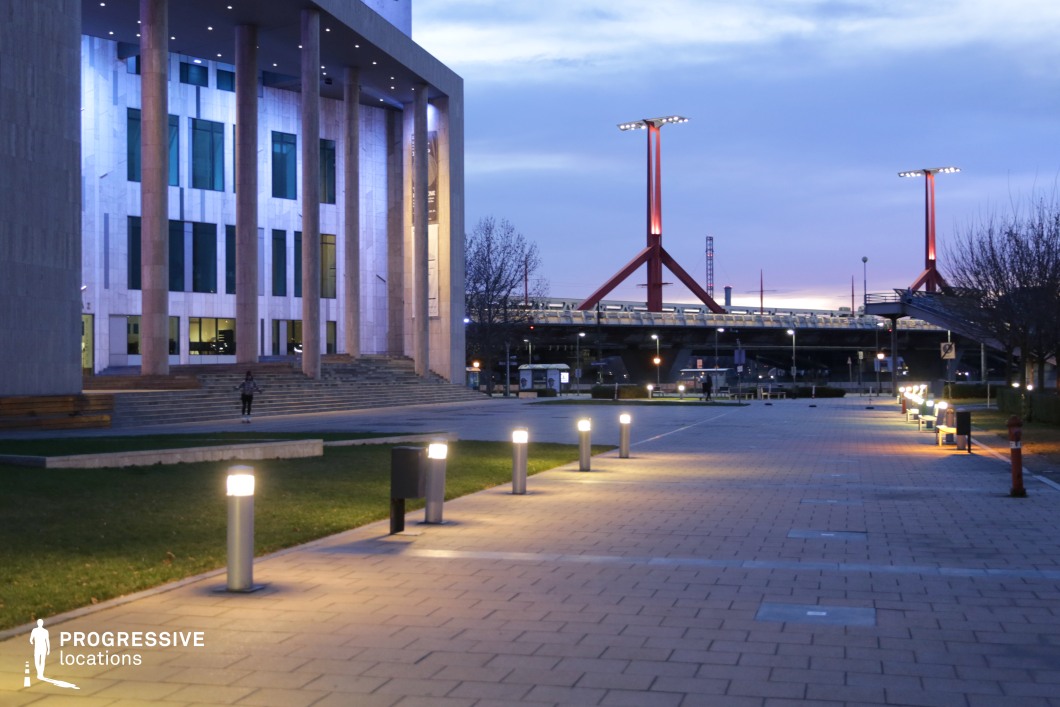 The image presents a contemporary urban plaza with striking architectural elements. The view is dominated by a large building with a grand columned entrance, reflecting a monumental, artistic style. The lighting is well-designed, with fixtures illuminating the pathway and surrounding areas, enhancing the aesthetic appeal of the space. In the background, a modern bridge structure with tall, red-painted support pillars adds an industrial touch, contrasting with the sleek and clean lines of the building. This location is ideal for urban scene production, offering a mix of classical elegance and modern, functional design. The well-lit public space and the bold design elements provide a dynamic environment for film and commercial shoots.