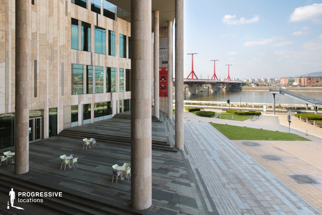 This image showcases an expansive modern plaza in front of an impressive marble-clad building, highlighted by towering columns and wide, ascending steps. The building's large glass windows reflect the surrounding environment, allowing natural light to flood the space. At the base of the structure, outdoor seating areas are set against the backdrop of a pedestrian-friendly space. The scene is framed by a striking red-painted bridge spanning the river, creating a dynamic contrast with the natural green areas and the smooth stone surfaces. This open, well-lit public space offers an ideal location for filming urban scenes, with its elegant architecture and inviting atmosphere. The combination of modern design and scenic river views adds to its cinematic potential.