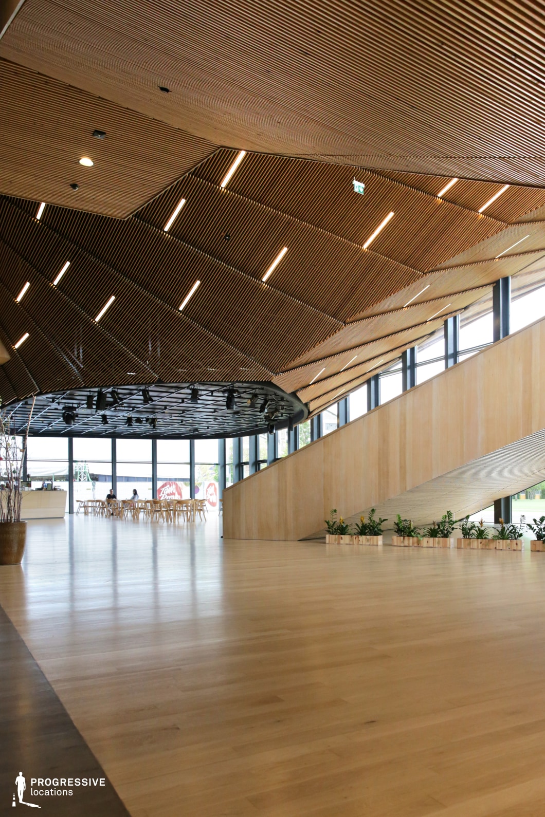 This image features a spacious interior with an inviting, modern design. The ceiling is characterized by sleek wooden panels, arranged in a dynamic angular pattern, with integrated lighting that enhances the room's ambiance. Large windows line the walls, providing ample natural light and views of the exterior. A wide wooden staircase gently rises from the lower level, its clean lines blending seamlessly with the minimalist interior. The room is furnished with simple, contemporary furniture, including dining tables and potted plants that add a touch of greenery. The use of natural wood finishes and warm tones creates a calming, sophisticated atmosphere, making this space suitable for various film scenes that require a clean, modern aesthetic with a focus on natural materials.