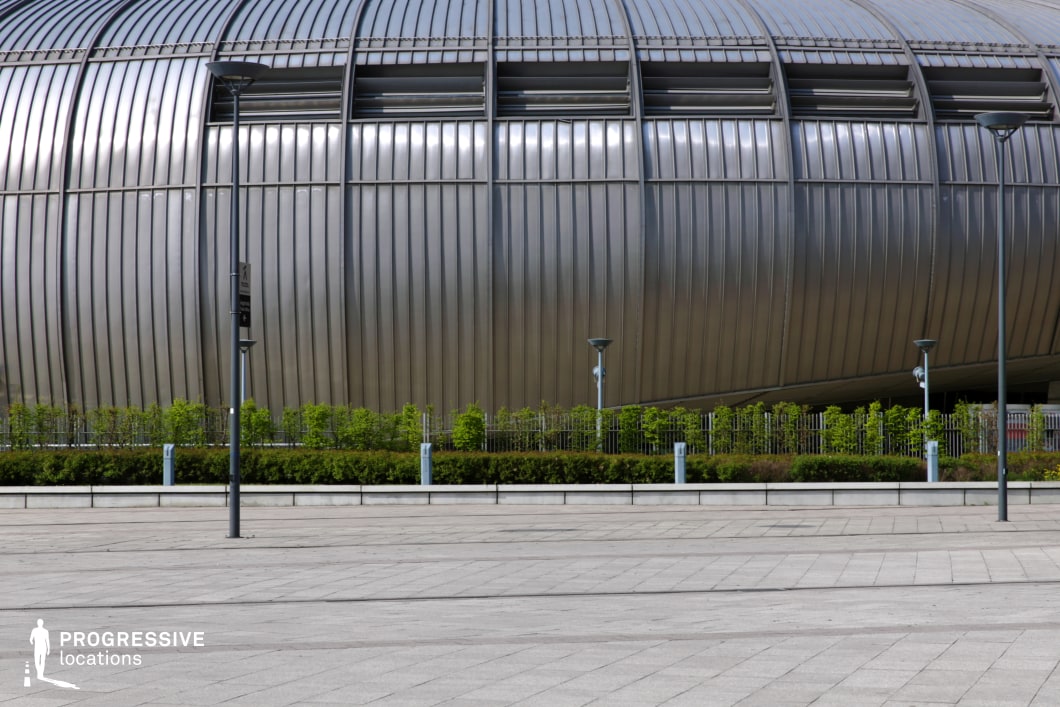This image presents the striking exterior of a modern event hall with a bold, metallic facade. The building’s curved and ribbed design creates a sleek, industrial appearance, dominated by reflective metal panels. The structure’s futuristic aesthetic is enhanced by strategically placed ventilation slats along its surface. In front of the building, a well-maintained garden with neat hedges and small trees lines the base, creating a contrast between the rigid geometry of the architecture and the soft greenery. The open plaza in front is paved with uniform gray tiles, providing a clean and expansive setting ideal for film productions, offering a dynamic visual of modern urban architecture with an industrial flair. This location would work well for scenes that require a cutting-edge, bold environment.
