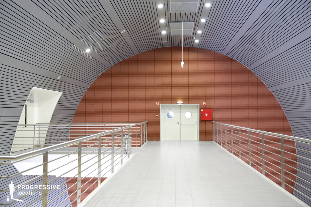 This futuristic corridor features a sleek, modern aesthetic with its prominent curved ceiling and wall panels, which are arranged in a linear, striped pattern. The ceiling design showcases a series of alternating light panels that enhance the feeling of spaciousness, while the red brick wall provides a striking contrast to the otherwise neutral tones. A stainless steel railing runs along the edge of the walkway, adding a polished and functional detail to the space. A round, porthole-like door in the center creates a sense of intrigue and futuristic design, complemented by subtle, recessed lighting that highlights the architectural features. The corridor's minimalist approach with industrial elements makes it an ideal choice for sci-fi or high-tech themed scenes, perfect for futuristic or corporate settings in film productions. The clean lines and vibrant colors also offer opportunities for dynamic visual contrasts in storytelling.