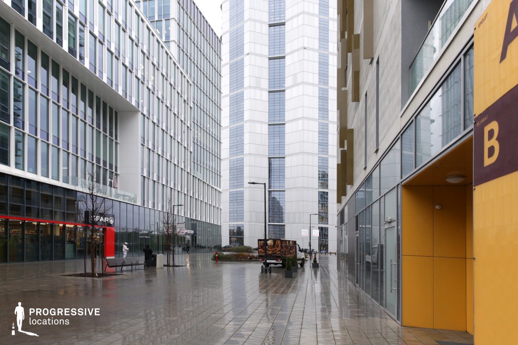 This modern urban scene features a sleek, glass-fronted building dominating the street. The polished pavement reflects the surrounding architecture, adding a dramatic effect to the environment. On the left, a store with a red and white sign stands in stark contrast to the sleek glass surfaces of the nearby skyscraper. The right side of the frame reveals a yellow-painted door and contemporary, minimalist design details on the adjacent building, suggesting a corporate or commercial atmosphere. The street exudes a clean, functional aesthetic, with the sharp architectural lines of the skyscraper combining reflective glass and geometric edges that define the modern city block. The polished finish of the pavement, paired with the soft lighting, creates a moody, cinematic feel, ideal for scenes depicting urban sophistication, business districts, or a reflective atmosphere in film productions. The space is suitable for various types of shots, from street-level interaction to high-angle captures of the surrounding skyline.