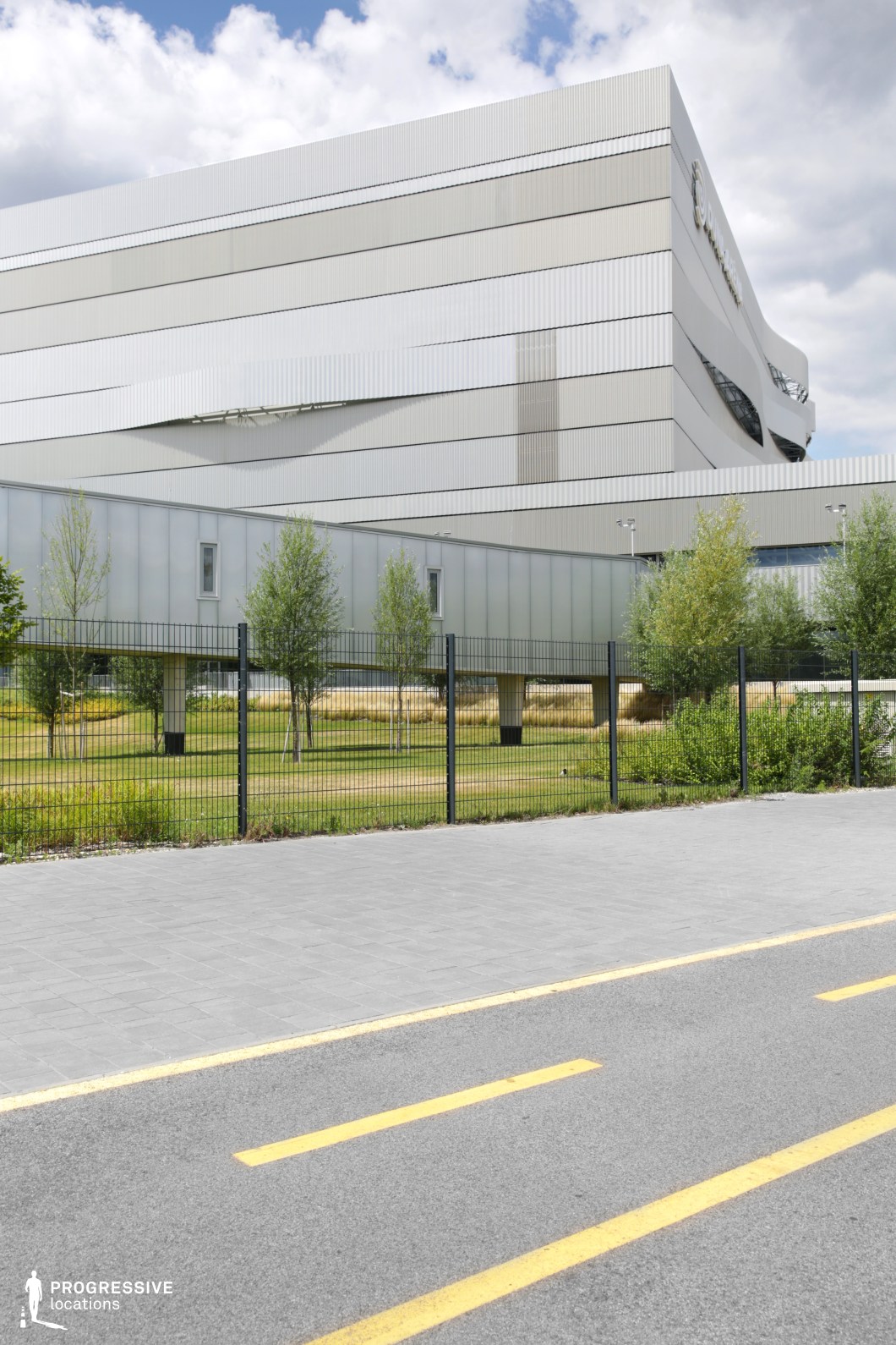 This modern sports facility is framed by a sleek metallic exterior that suggests a contemporary, industrial design. The building, with its vertical lines and smooth surface, contrasts beautifully with the surrounding greenery and well-maintained outdoor space. In the foreground, a paved bike track is marked with bold yellow lines, creating a clear path that leads toward the building. A black fence encloses the perimeter, offering security while maintaining the minimalist and functional aesthetics of the space. The lawn beyond the fence is dotted with trees, adding a natural element to the urban setting. This clean, open environment is ideal for scenes that require a dynamic, athletic backdrop or a futuristic urban setting. The simplicity and scale of the space can complement a variety of film genres, from sports dramas to corporate or tech-focused productions.