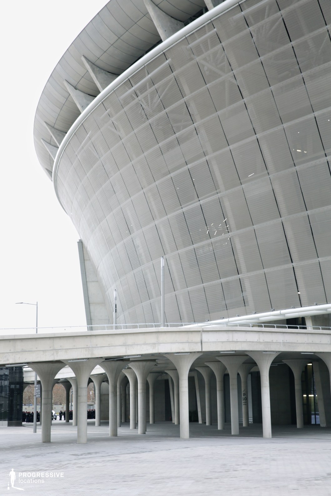 This modern stadium design features a sweeping, curved facade with a distinctive grid pattern that creates an industrial, cutting-edge aesthetic. The exterior is supported by massive, concrete pillars, which stand tall and provide structural support for the upper levels of the building. The gray concrete architecture contrasts beautifully with the surrounding paved area, which is open and clean, offering ample space for movement. The stark and minimalist style of the exterior, combined with the strong geometric lines, makes this location suitable for dynamic, modern film productions, particularly those requiring an urban or industrial backdrop. The openness of the plaza beneath the stadium is ideal for wide shots, providing versatility for various cinematic uses, from sports-themed scenes to futuristic cityscapes.