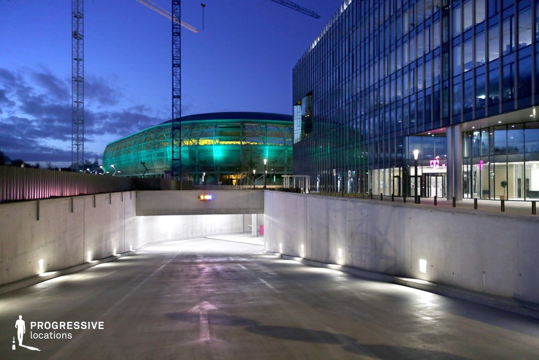 This nighttime shot captures a modern underground parking area adjacent to a large stadium. The industrial concrete structure leads into a dimly lit parking lot with directional lighting on the floor, creating a sleek, functional atmosphere. In the background, the illuminated facade of the stadium glows with a distinct green hue, adding a dynamic contrast to the otherwise neutral tones of the surrounding environment. The surrounding glass building and construction cranes in the background reflect the contemporary architectural style of the area. The setting offers a versatile location for filming scenes requiring an urban, futuristic, or underground aesthetic, with the stadium's glowing exterior offering an interesting focal point.