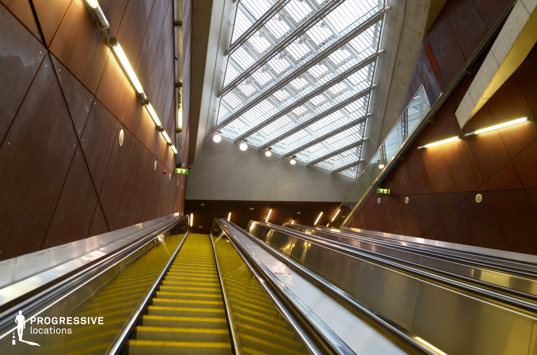 The image captures a modern escalator inside a sleek, industrial space. The escalator features bright yellow steps, contrasting with the polished stainless steel railings and providing a striking visual effect. The surrounding walls are clad in rust-colored metal panels, adding an industrial aesthetic. Above, a large skylight allows natural light to flood the space, illuminating the escalator and emphasizing the clean lines of the architecture. The interior lighting, with strategically placed LED lights, highlights the angular features of the walls, further accentuating the design of the space. This modern, industrial setting is well-suited for film productions that require a contemporary, visually dynamic environment, combining sharp contrasts with a clean, minimalistic design.