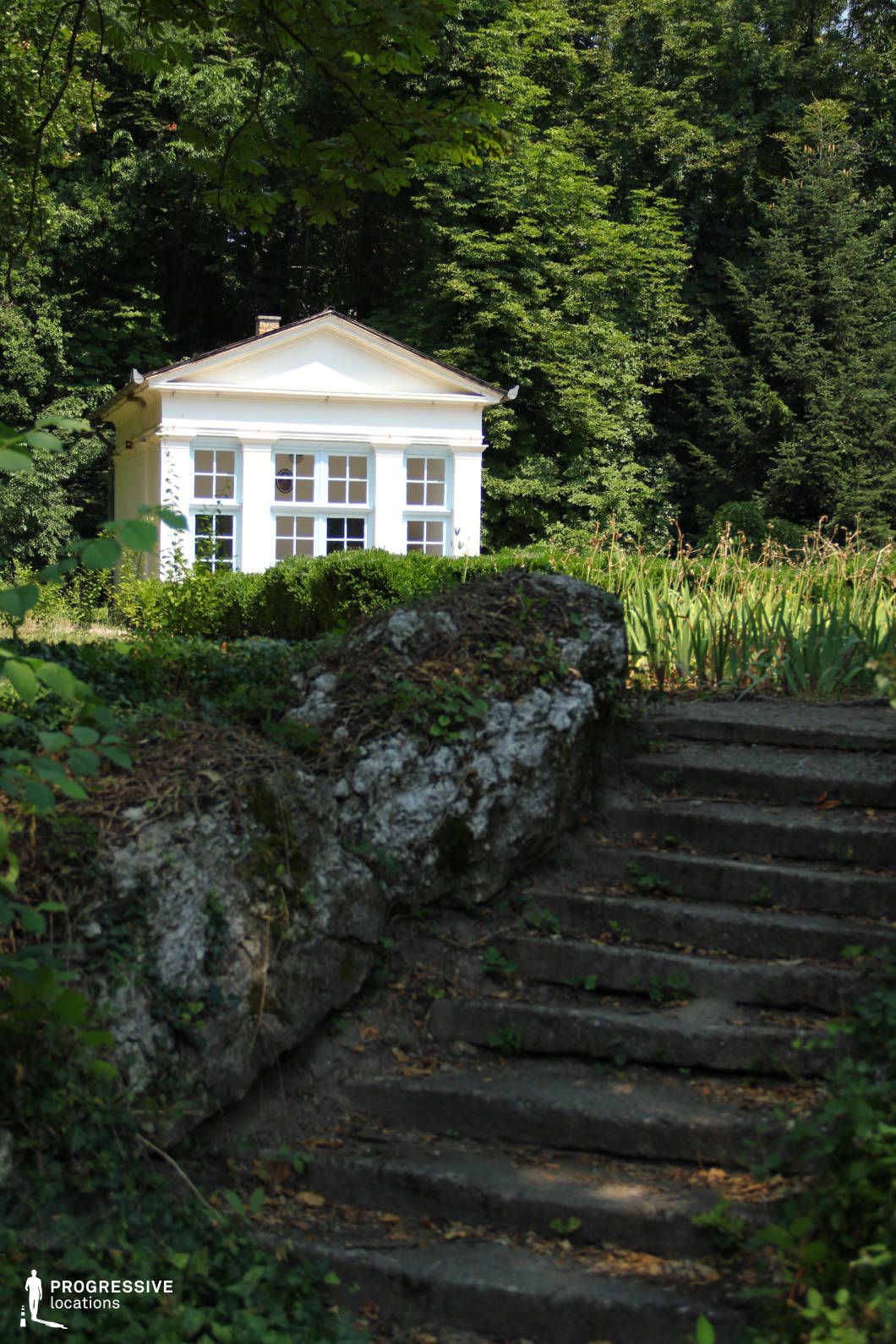 This peaceful image depicts a white classical pavilion nestled in the greenery of a park. The building is framed by lush trees, adding a sense of privacy and serenity. The structure itself has a refined, neoclassical design with large windows, creating a harmonious contrast with the dense foliage surrounding it. In the foreground, a set of stone steps leads up to the pavilion, partially overgrown with ivy, adding a natural touch to the scene. The well-maintained bushes and garden plants around the pavilion enhance the feeling of elegance and tranquility. This location offers a timeless, elegant setting, ideal for historical or intimate scenes in film or photography. The location's secluded nature, coupled with its refined architecture, makes it perfect for portraying a quiet retreat or a place of contemplation. The combination of natural elements and classical design offers flexibility for various creative uses, from romantic backdrops to sophisticated moments.