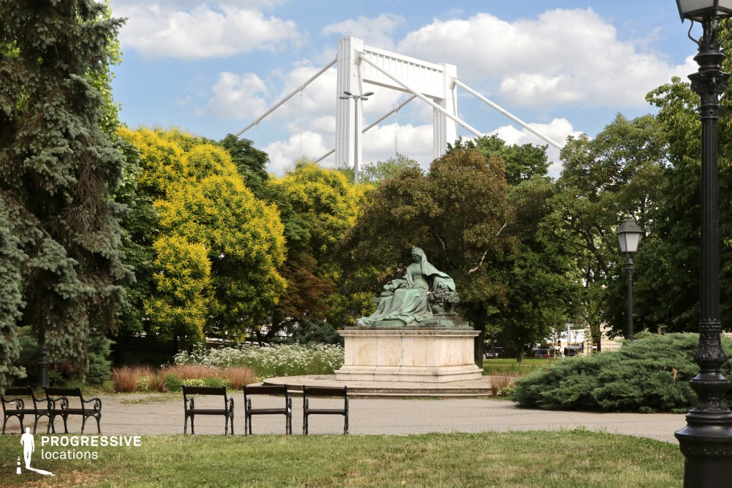 This vibrant park scene captures a beautiful blend of nature, art, and modern architecture. In the foreground, several benches are lined up near a bronze statue, which is set on a raised platform. The statue, detailed and artistic, adds a focal point amidst the lush greenery. Behind the statue, trees in varying shades of green create a natural frame, with bright yellow accents in the foliage providing a touch of vibrancy. A modern white bridge towers in the background, creating a striking contrast with the traditional park elements. The combination of the park’s natural beauty, the classical sculpture, and the contemporary bridge offers a dynamic visual experience. This location provides excellent potential for film scenes that require both historical elegance and modernity, making it suitable for a variety of narratives. The clean lines of the bridge and the serene atmosphere of the park make this site ideal for thoughtful, reflective moments, or scenes that need a backdrop with a mix of both the old and new.