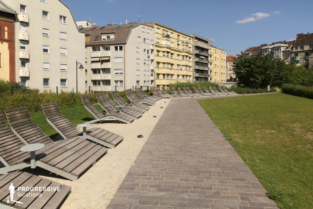 This image captures a well-designed urban relaxation space featuring a row of modern wooden sun loungers arranged along a curved gravel pathway. The loungers, equipped with small metal side tables, offer a functional and stylish outdoor seating option for public use. A paved walkway made of rectangular stone tiles leads through the area, providing easy accessibility. In the background, a mix of residential apartment buildings with diverse architectural styles, ranging from classic to modern, frames the scene. The facades exhibit neutral tones, with some balconies adding character to the urban setting. The surrounding greenery, including neatly trimmed grass and low shrubs, balances the built environment, creating a tranquil atmosphere within the city. This location is ideal for lifestyle photography, urban relaxation themes, or showcasing contemporary public space design with a focus on comfort and accessibility.