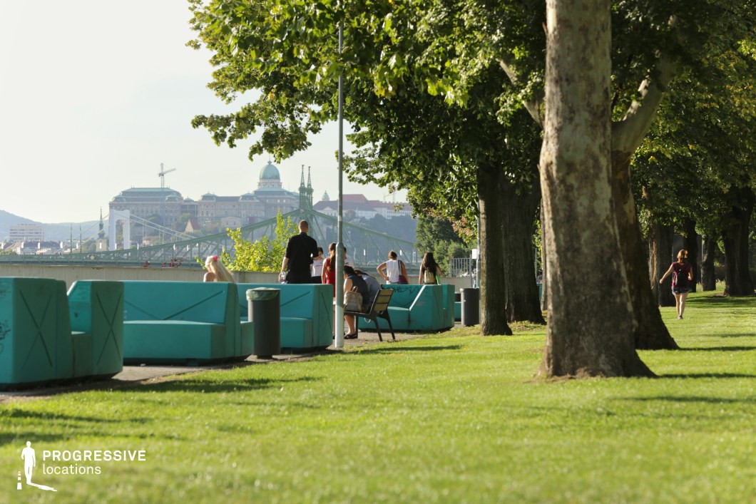 This image presents a scenic riverside promenade lined with tall, mature trees providing ample shade. The well-manicured grass and wide pedestrian walkway create a welcoming and open atmosphere. In the foreground, uniquely designed turquoise concrete seating elements add a modern and vibrant touch to the urban landscape. A wooden bench with a metal frame offers a comfortable resting spot along the path. The background features a striking city skyline with historic architecture, including a prominent domed building and bridges spanning the river, reinforcing the location’s cultural and architectural richness. The lush greenery contrasts with the urban environment, making this setting ideal for relaxation, social interactions, or scenic strolls. This location is perfect for travel and lifestyle photography, cityscape imagery, or productions emphasizing the blend of nature and historic urban environments.