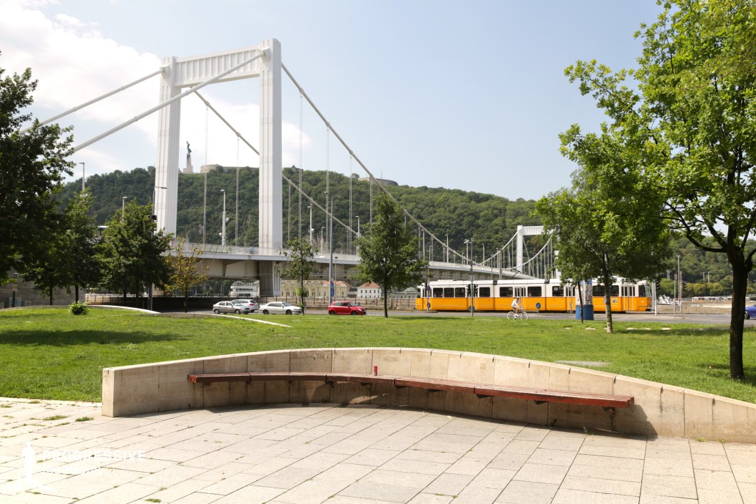 This image showcases an open urban plaza with a curved concrete bench in the foreground, set against the backdrop of a striking suspension bridge. The plaza is paved with large stone tiles, and the surrounding green spaces are dotted with trees, adding a natural contrast to the structured environment. In the middle ground, a classic yellow tram runs parallel to the bridge, contributing to the dynamic city atmosphere. The suspension bridge, with its clean white cables and towering pylons, spans the river, connecting different parts of the city. In the background, a lush green hillside provides a natural element, enhancing the contrast between urban infrastructure and natural surroundings. This location is ideal for showcasing public transportation, cityscape compositions, or architectural photography. Its open space and iconic urban features make it a versatile setting for various creative projects.