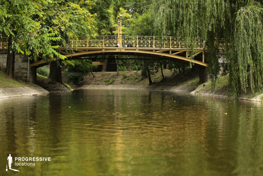 This image captures a serene natural setting featuring a decorative pedestrian bridge spanning a calm body of water. The bridge, painted in a soft golden hue, showcases intricate metal railings and a vintage-style lamppost at its center, adding a touch of elegance to the scene. Its arched design blends harmoniously with the lush greenery surrounding it. The waterway reflects the surrounding trees, creating a peaceful, mirror-like effect. Overhanging willow branches frame the composition, adding depth and softness to the visual. The embankment is lined with stone and grass, emphasizing the natural and well-maintained landscape. This location is ideal for romantic, tranquil, or historic-themed visuals. Its picturesque qualities make it a versatile setting for period pieces, nature cinematography, or elegant lifestyle photography.