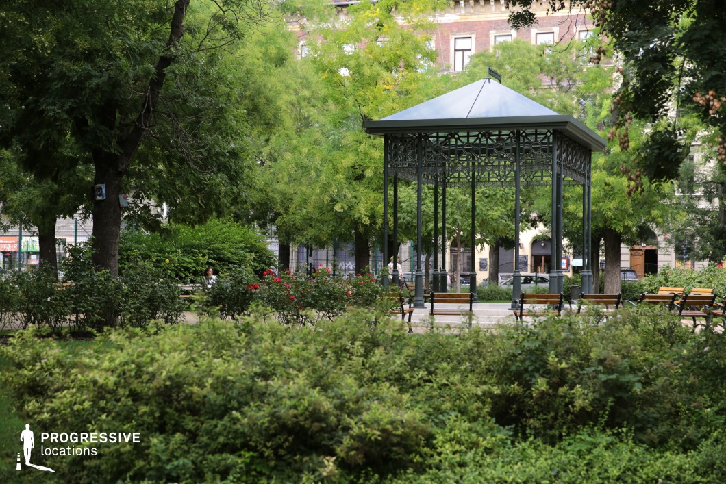 This image captures a charming urban park with lush greenery and a beautifully designed metal gazebo as its focal point. The gazebo features intricate wrought-iron detailing and a classic pitched roof, giving it an elegant, vintage appeal. Surrounding the structure, wooden benches with metal frames provide ample seating, while neatly trimmed rose bushes and various plants add color and vibrancy to the setting. Tall, mature trees create a natural canopy, offering shade and enhancing the park's tranquil atmosphere. In the background, historic buildings with classic architectural elements contribute to the sense of timelessness and sophistication. The park appears well-maintained, making it an inviting location for relaxation, conversation, or quiet contemplation. This location is ideal for period-piece photography, romantic or literary-inspired visuals, or productions that require an intimate and picturesque urban setting.