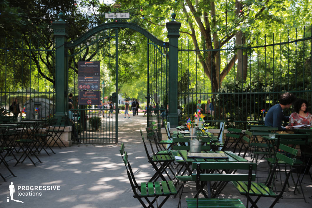 This image captures a picturesque café nestled at the entrance of a historic city park. A tall, elegant wrought-iron gate, painted deep green, serves as a welcoming focal point, opening into a sunlit park with lush trees and inviting pathways. The gate is adorned with decorative elements and signage, reinforcing the location’s historic and cultured ambiance. In the foreground, the café terrace features classic green metal tables and chairs arranged in an inviting manner. Small flower vases and decorative pinwheels add a playful and welcoming touch to the tables. The seating area is shaded by large trees, creating a dappled light effect on the ground, enhancing the cozy and relaxed atmosphere. Beyond the gate, the park’s vibrant greenery and neatly arranged flower beds provide a beautiful natural backdrop. The combination of the historic gate, inviting café, and lush park setting makes this an ideal location for lifestyle photography, outdoor dining promotions, or cinematic scenes centered around urban leisure and relaxation.