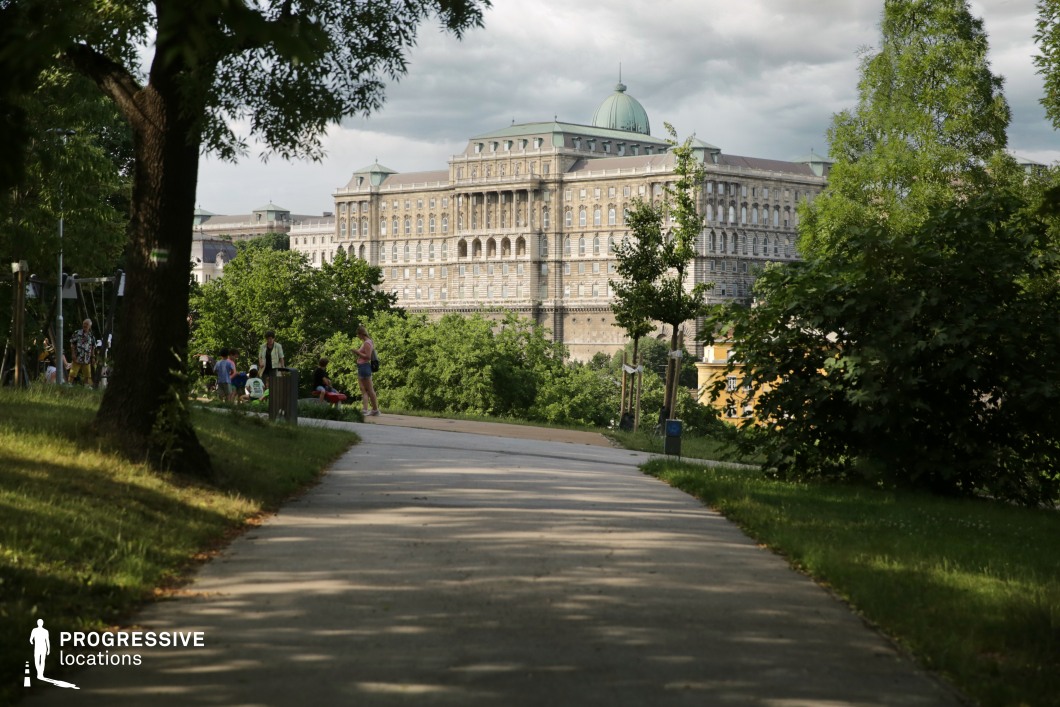 This scenic view captures a wide, paved pathway gently sloping through a lush park, leading toward the grand Buda Castle perched above. The foreground is lush with greenery, with trees and grass flanking the path, creating a peaceful, open atmosphere. In the distance, the imposing Buda Castle dominates the skyline, with its large, historical architecture contrasting beautifully with the natural surroundings. The well-maintained path provides a sense of flow, guiding the viewer’s eye toward the majestic castle. This location is perfect for cinematic scenes that require a mixture of serene parkland and dramatic, historic architecture. The combination of the park’s tranquility and the grandeur of Buda Castle makes this an ideal setting for both wide-angle landscape shots and focused close-ups capturing the intricate details of the castle's facade. The openness of the space and its proximity to the city offer versatility for various creative projects.