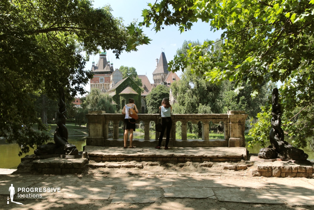 This image showcases a picturesque stone lookout overlooking a tranquil park and an ornate, fairy-tale-like castle in the background. The foreground is framed by lush green foliage, with dappled sunlight filtering through the leaves, casting soft shadows on the aged stone pavement. The balustrade, made of weathered stone, adds an old-world charm to the scene, while decorative sculptures on either side contribute to the historical atmosphere. Beyond the lookout, the castle features steeply pitched roofs, turrets, and ivy-covered facades, blending Gothic and Renaissance architectural influences. The surrounding landscape is filled with tall trees, weeping willows, and a gently flowing body of water, creating a serene and romantic ambiance. This location is ideal for period-piece cinematography, fantasy-themed photography, or historical storytelling. The combination of architectural grandeur and natural beauty makes it a visually captivating setting for creative projects.