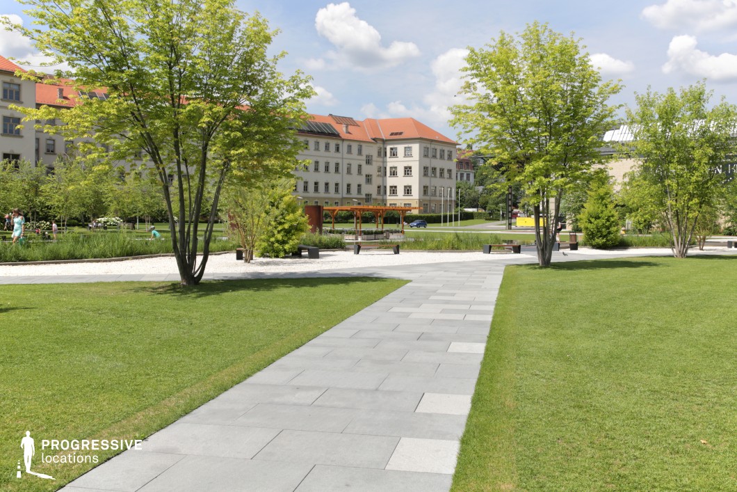 This image showcases a meticulously designed urban park featuring a contemporary layout with clean lines and structured landscaping. A sleek, light-gray paved walkway leads through the open green space, bordered by young trees with vibrant foliage. The neatly trimmed grass and carefully arranged vegetation contribute to the park’s well-maintained and inviting atmosphere. In the background, classical buildings with symmetrical facades and red-tiled roofs provide an elegant contrast to the modern park design. A wooden pergola with seating adds a functional and aesthetic focal point, while minimalist benches and white gravel pathways enhance the park’s structured yet natural appeal. This location is ideal for architectural photography, urban lifestyle imagery, or projects focusing on the integration of green spaces in city environments. The balance of classical and contemporary elements makes it a visually appealing setting for creative productions.