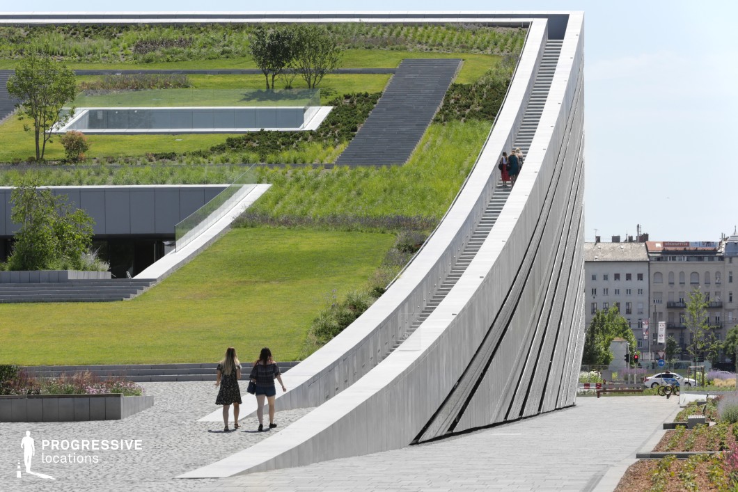 This image showcases an innovative architectural structure featuring a sloping, multi-tiered green roof that seamlessly blends with the surrounding landscape. The design incorporates extensive vegetation, neatly arranged grass terraces, and reflective glass railings, creating a visually striking fusion of nature and modernity. A wide pedestrian ramp extends along the curvature of the building, allowing for an elevated perspective and interaction with the space. The geometric lines of the building, combined with the lush greenery, contribute to a sleek, eco-conscious urban aesthetic. In the background, traditional buildings contrast with the contemporary design, emphasizing the architectural innovation of the space. The foreground features a cobblestone pathway leading towards the structure, enhancing accessibility and integration with the environment. This location is ideal for urban landscape photography, futuristic or eco-friendly-themed productions, and architectural showcases focusing on sustainable design. Its dynamic form and integrated natural elements make it a visually compelling and adaptable setting.