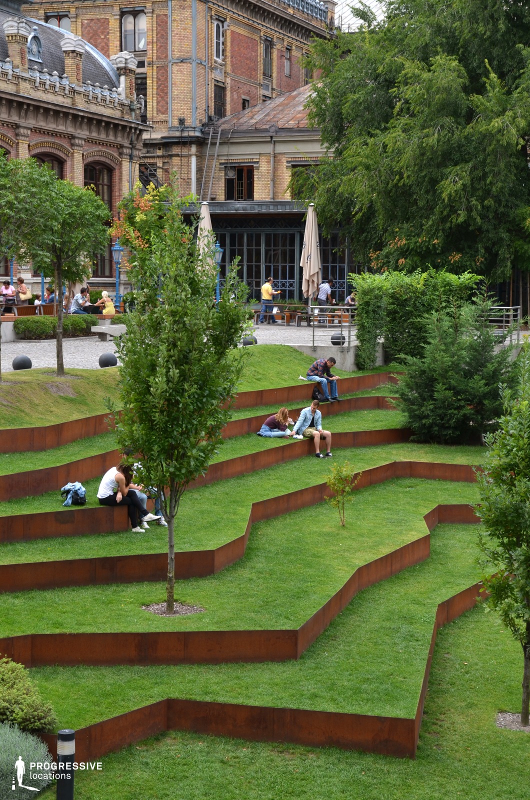This image showcases a unique urban park with a terraced landscape design featuring angular, rust-colored metal borders that define the stepped grassy seating areas. The geometric arrangement of these terraces creates a visually dynamic contrast against the lush green lawn, offering a modern and artistic take on outdoor seating. Young trees and shrubs are strategically placed throughout, enhancing the natural ambiance while maintaining an open, structured layout. In the background, an ornate historic building with intricate brickwork, arched windows, and decorative rooftop elements serves as a striking architectural contrast to the park’s contemporary design. A café with large umbrellas and blue metal accents sits adjacent to the park, blending urban leisure with green public space. The mix of modern landscaping and classical architecture makes this location a visually engaging setting. This park is ideal for urban design showcases, architectural photography, and productions focusing on the fusion of historic and contemporary elements in public spaces. The structured yet organic flow of the terraces provides depth and versatility for creative compositions.