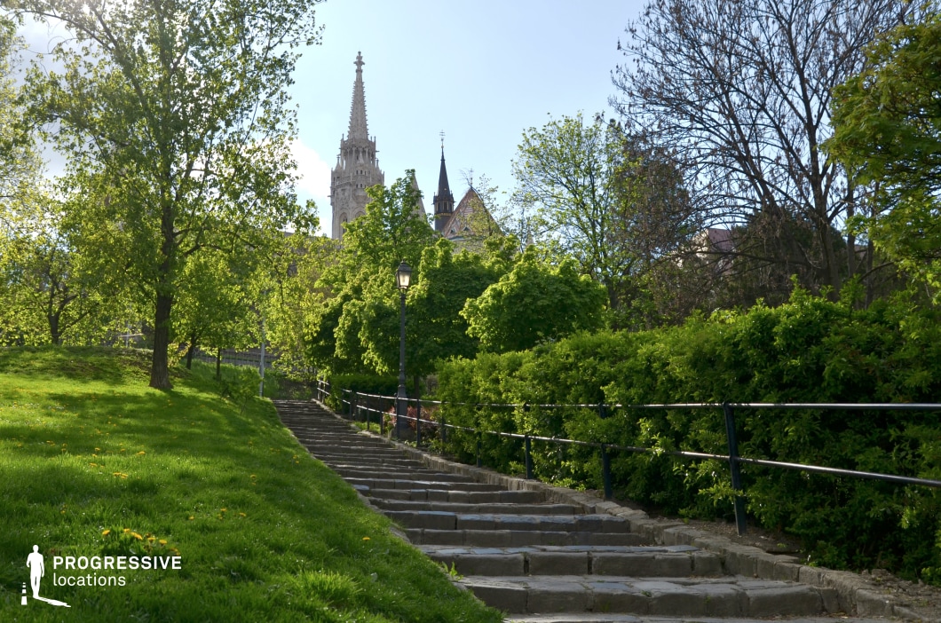This image captures a picturesque stone stairway leading up to a historic architectural landmark partially visible in the background. The stairway, bordered by a black metal railing, is lined with vibrant green hedges and mature trees, creating a natural corridor that enhances the scene’s depth and perspective. The lush grass slopes gently alongside the stairs, dotted with small yellow flowers that add to the inviting atmosphere. A vintage-style lamppost stands along the path, reinforcing the timeless character of the location. The background features an intricate Gothic-style spire and colorful tiled rooftops, hinting at a historic and culturally significant site. The combination of greenery and architectural elements provides a balanced and visually appealing composition. This location is ideal for cinematic establishing shots, travel photography, or projects highlighting historic urban landscapes. The blend of natural beauty and architectural grandeur creates a versatile and evocative setting for creative productions.