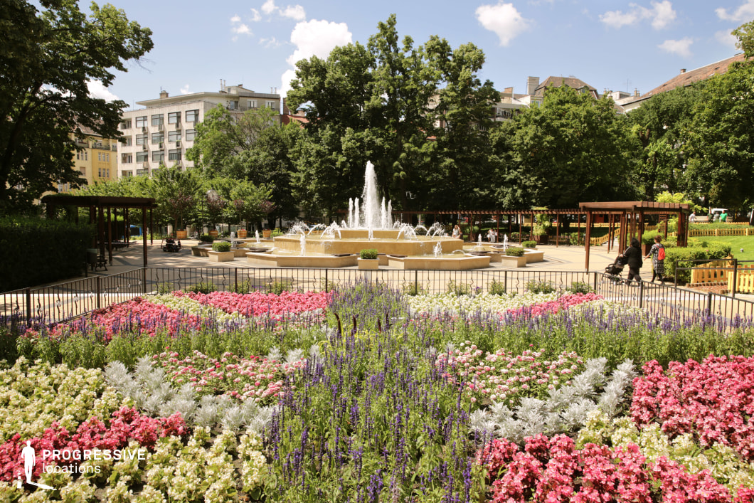 This image showcases a beautifully landscaped urban park featuring a grand multi-tiered fountain as its central focal point. The cascading water jets add movement and vibrancy, while the surrounding symmetrical flower beds, filled with a variety of colorful blooms, create a visually striking foreground. A black wrought-iron fence elegantly frames the garden, enhancing its structured aesthetic. Lush green trees provide shade and contrast with the bright floral arrangements. Wooden pergolas and benches are strategically placed throughout the park, offering inviting resting spots. The surrounding architecture, featuring a mix of modern and historic buildings, integrates the park into the urban fabric while preserving a sense of tranquility and elegance. This location is ideal for urban lifestyle photography, cinematic park scenes, or productions requiring a refined and picturesque outdoor setting. The combination of classical design elements, vibrant nature, and structured landscaping makes it a versatile and visually compelling environment.