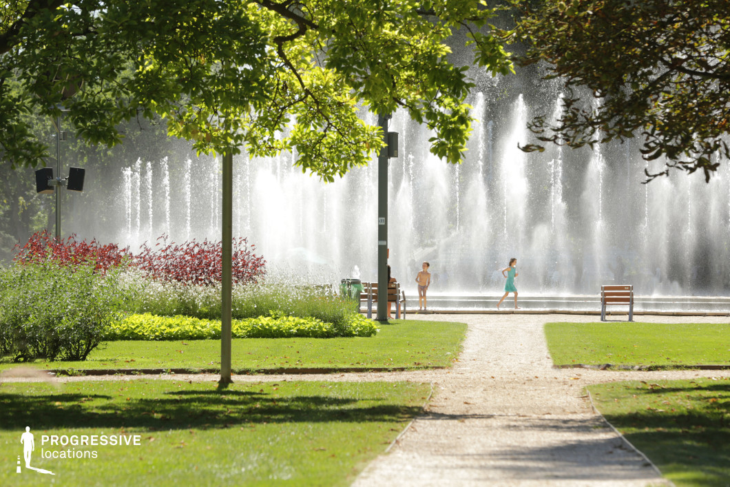 This image captures a stunning park scene featuring a large, dynamic water fountain as the central element. The cascading water jets create a rhythmic display, adding a sense of movement and refreshing energy to the setting. Surrounding the fountain, a variety of colorful plants and well-manicured lawns enhance the natural beauty of the space. A gravel pathway leads towards the fountain, flanked by lush greenery, red-leafed bushes, and strategically placed wooden benches. Tall trees provide natural shade, casting dappled light onto the grass and walkways. The structured landscaping, combined with the openness of the park, makes this location an ideal setting for peaceful relaxation or scenic urban visuals. This park is well-suited for cinematic sequences, lifestyle photography, or commercial productions requiring an elegant outdoor space with a balance of natural and architectural elements. The striking combination of water, greenery, and open pathways offers a visually engaging and inviting atmosphere.
