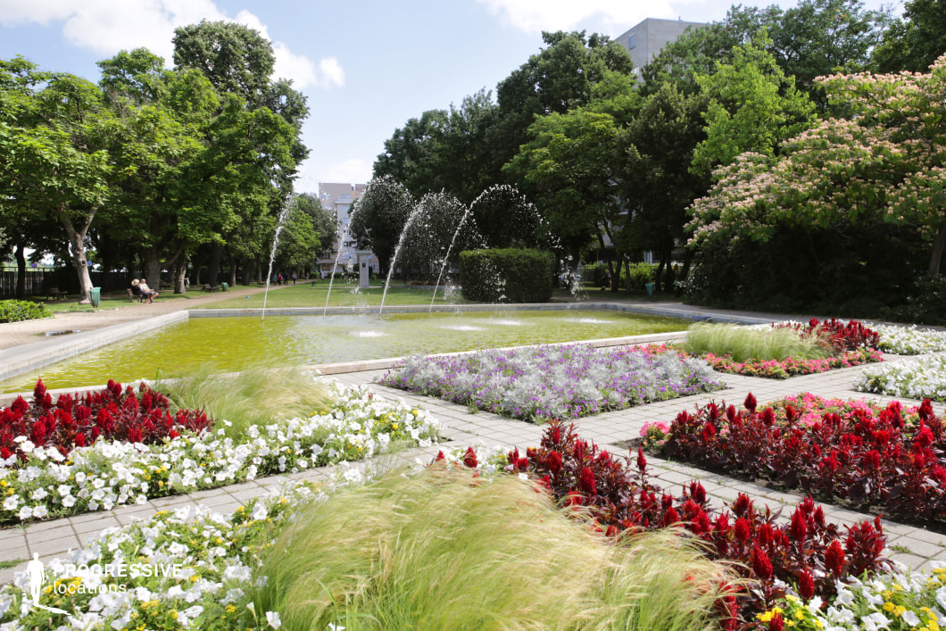 This lush urban park features a beautifully maintained water fountain at its center, with symmetrical jets of water arching over a reflective pool. Surrounding the fountain, a variety of colorful flower beds, including red, white, and purple blooms, add a dynamic contrast to the neatly paved walkways and green lawns. Mature trees provide a natural canopy, offering both structure and shade to the space. In the background, a tree-lined pathway extends into the park, framed by carefully landscaped greenery and flowering shrubs. The combination of hardscape and softscape elements creates a balanced setting that is ideal for outdoor relaxation, film shoots, or advertising backdrops. This location offers a visually engaging mix of water, greenery, and floral design, making it suitable for scenic urban landscapes or elegant garden-focused visuals.