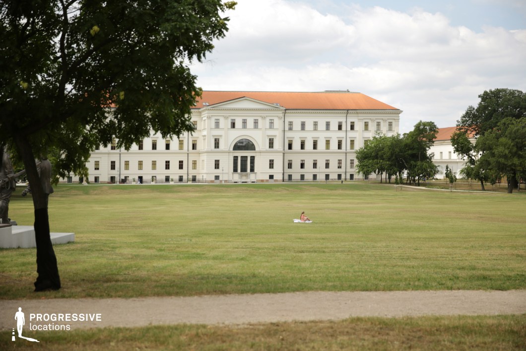  A vast open lawn leads up to a grand neoclassical building with a symmetrical facade, tall arched windows, and a prominent central entrance. The building's clean white exterior contrasts with its terracotta-tiled roof, lending it a stately presence. The foreground features a dirt pathway lined with scattered trees, their branches framing the scene and casting soft shadows. The well-maintained grass stretches across the entire space, creating an airy and tranquil atmosphere. The structured landscape, punctuated by a few evenly spaced trees, adds a sense of balance and openness. This location is ideal for historical or formal settings, period films, or scenes requiring an elegant and refined outdoor backdrop.