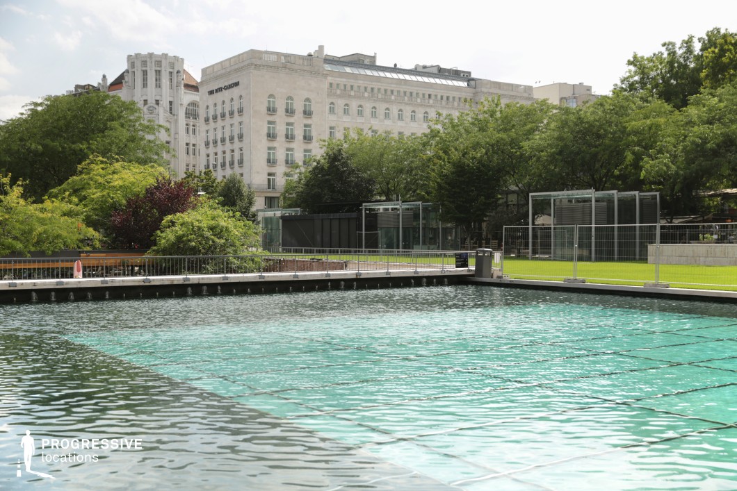 A large, clear water feature with a rippling surface is set within a well-manicured urban park. The area is bordered by a sleek metal railing and surrounded by lush green trees and neatly trimmed lawns, creating a balance between natural and modern elements. In the background, elegant historical and contemporary buildings rise above the landscape, their light-colored facades contrasting with the deep greenery. Glass-structured pavilions and fences add a contemporary touch to the space, suggesting a multifunctional area that could serve as a leisure or event venue. This location is ideal for sophisticated urban cinematography, promotional shoots, or tranquil outdoor scenes.